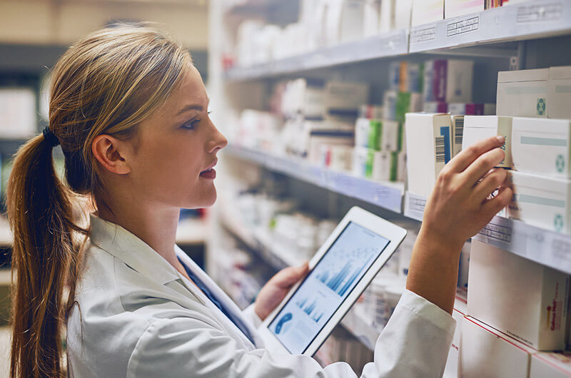 Pharmacist reviewing medication data on a digital device while managing prescriptions in a modern tech-enabled pharmacy workspace.