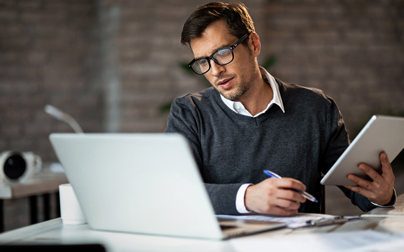 Businessman holding tablet device and pen while looking at laptop computer