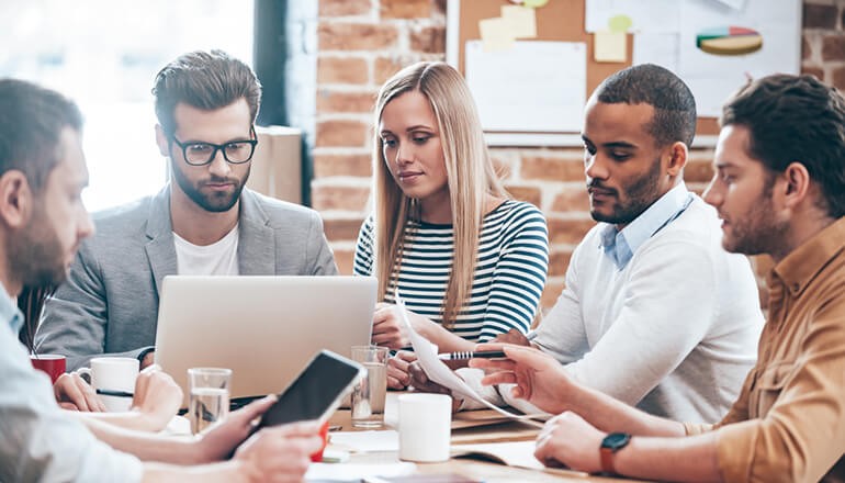 Group of business professionals gathered around desk with various devices