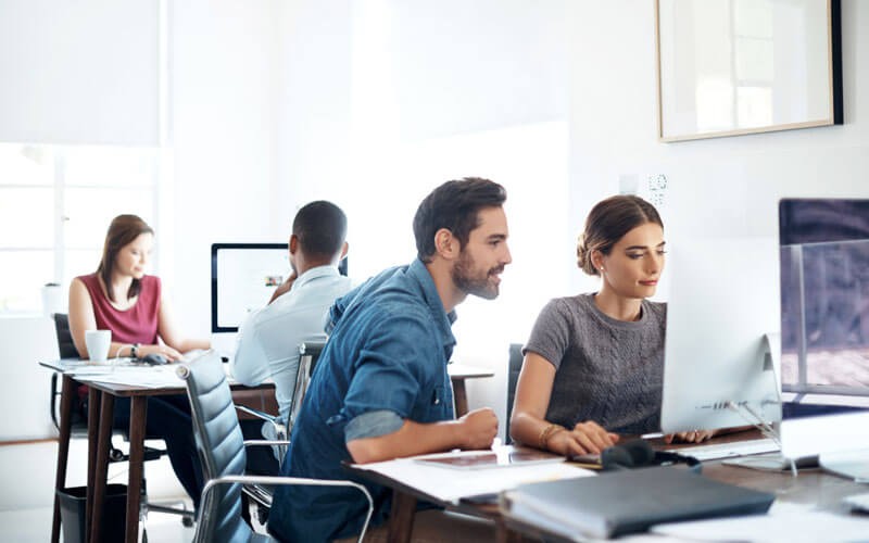 Two colleagues gather around desktop computer with team in background